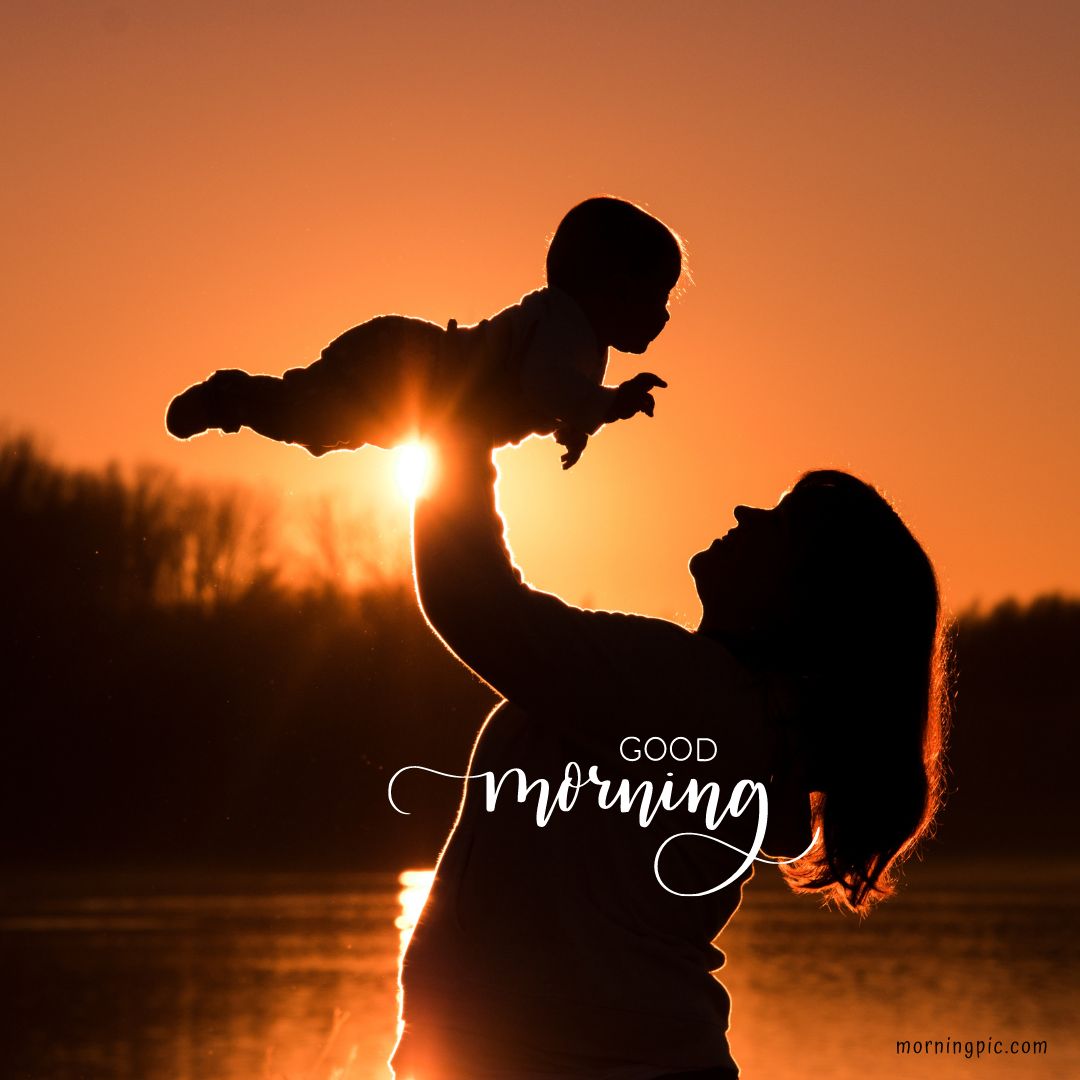 A silhouette of a person holding a baby up against a sunrise or sunset background near a body of water. The sky is radiant with warm hues. An artistic text overlay reads "Good Morning." The scene exudes warmth and tenderness, perfect for Good Morning Baby Images HD.