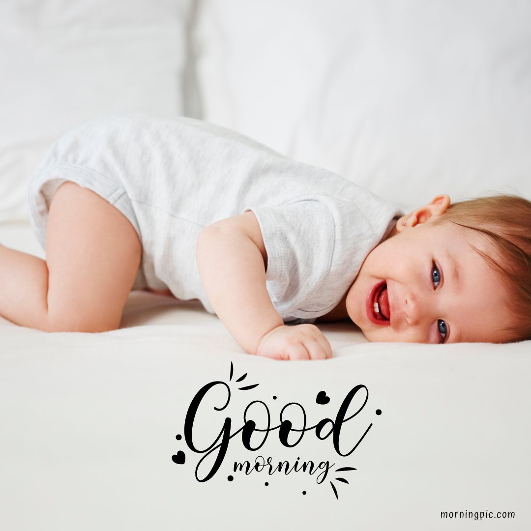 A smiling baby boy in a white onesie is lying on a white bed on their stomach, looking up with joy. The words "Good morning" in a decorative font are placed at the bottom of the image, making it one of the cutest Good Morning Images with Baby Boy.