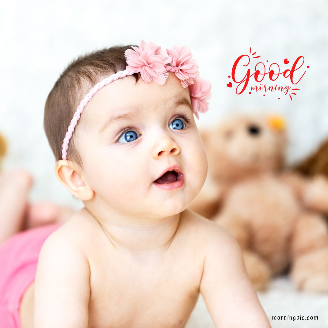 A baby girl with bright blue eyes and a pink headband adorned with flowers glances upwards, her mouth open in amazement. Blurred teddy bears form the background, while "Good morning" is written in red script on the top right corner. This captures the essence of good morning images with a baby girl.