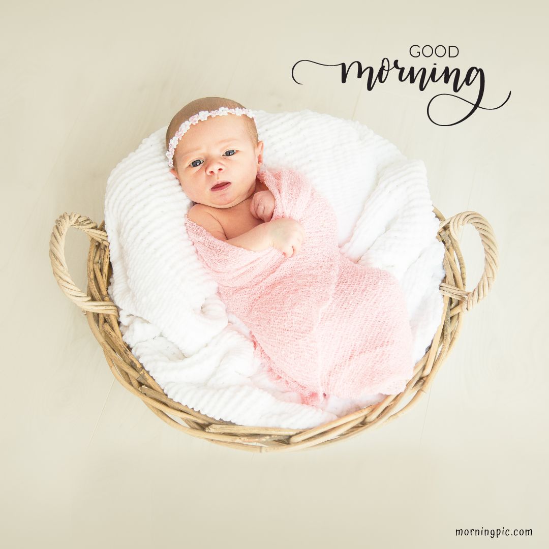 A baby wrapped in a pink blanket and wearing a white floral headband lies in a white cushioned basket. The words "Good Morning" are written above the baby in stylized script. The basket is placed on a light-colored floor. For more Good Morning Baby Images