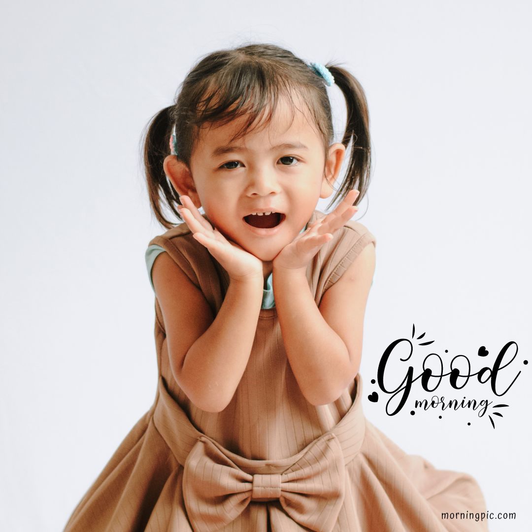 A young girl with pigtails, wearing a light brown dress with a bow, smiles with her hands framing her face. The text "Good Morning" is written in a decorative style beside her, perfect for those seeking Good Morning Baby Images.