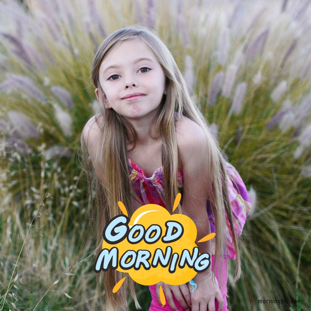 A young girl with long, straight hair is bending slightly forward, smiling at the camera while standing in front of tall, feathery grass. The words "GOOD MORNING" are displayed in bold, playful letters at the bottom of this charming Good Morning Baby Image.