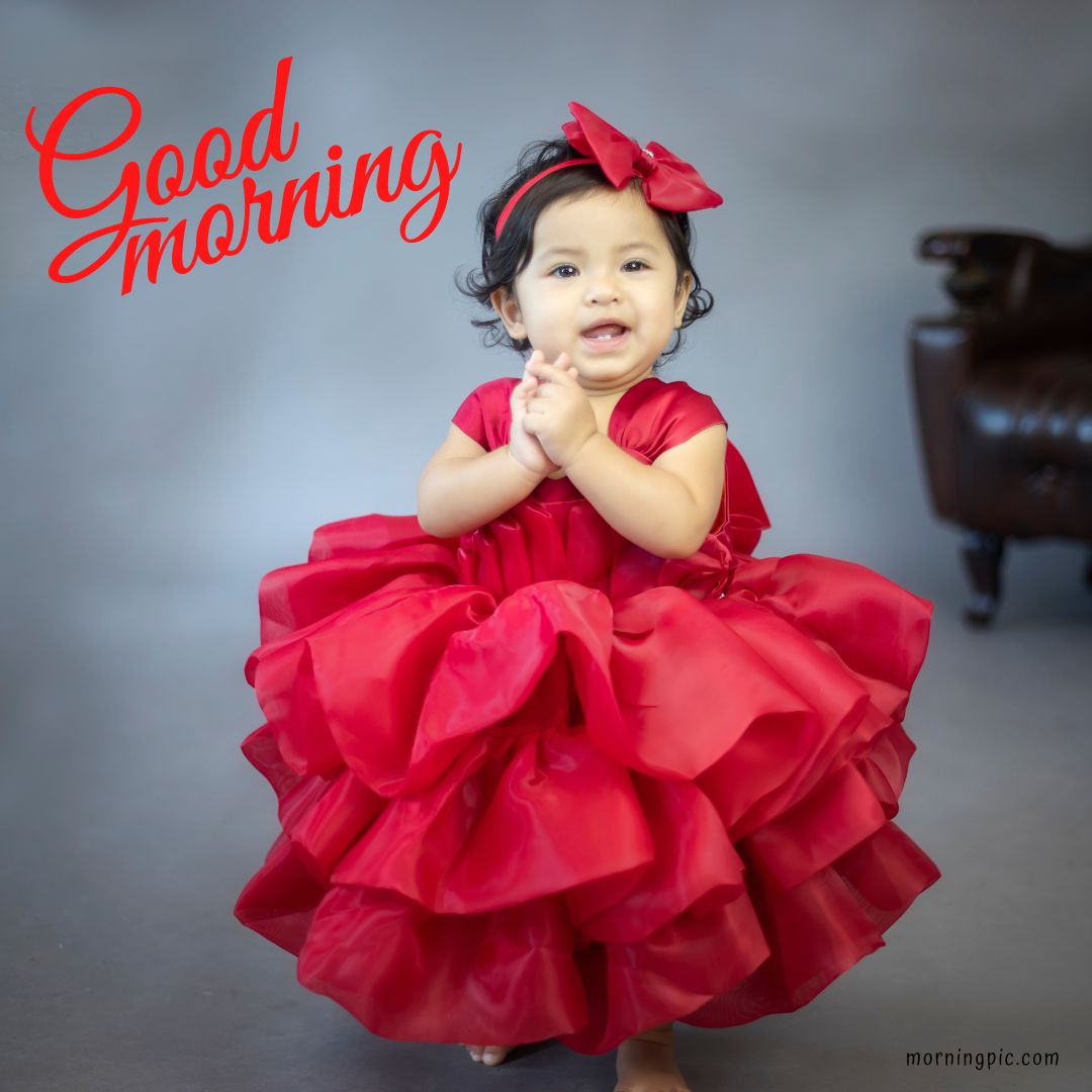 A young child wearing a vibrant red ruffled dress and red bow stands against a gray background. The child is smiling and has their hands clasped together. The text "Good Morning Baby Girl" is written in red at the top left corner of the image.