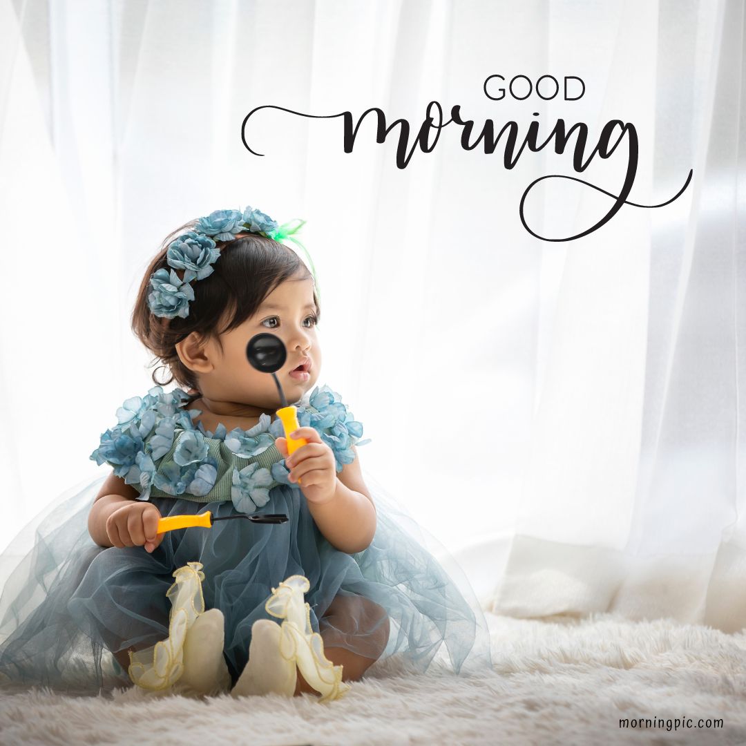 A small child in a fluffy blue dress and floral headband sits on a soft, white, fuzzy rug in front of sheer white curtains. The child holds a toy hammer and wrench and has a sticker on one cheek. This Good Morning Baby Girl image features text overlay that says, "Good morning.