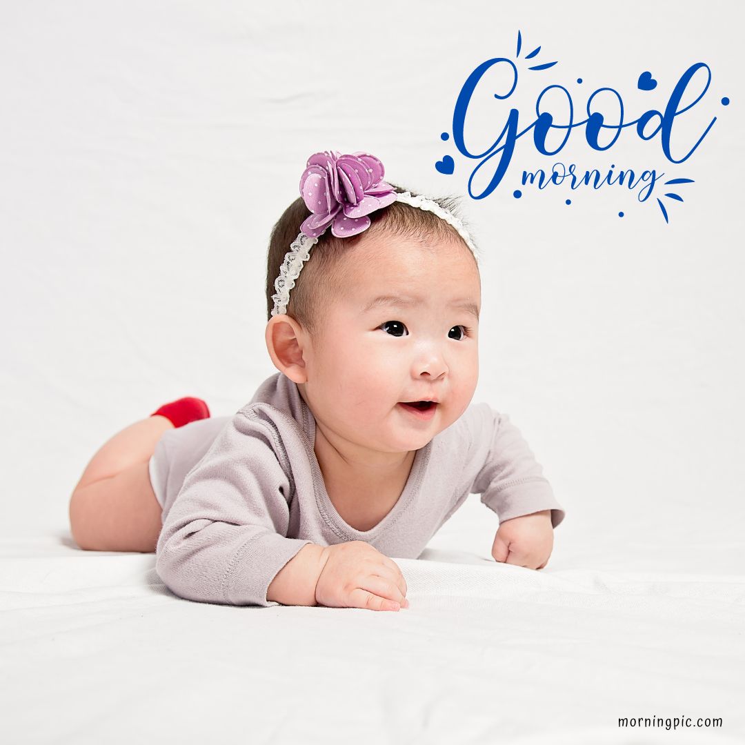 A baby girl wearing a light purple onesie and a headband with a purple flower lies on a white surface, smiling. The background is white with text that reads "Good Morning Baby Girl" in blue, accompanied by small blue hearts.