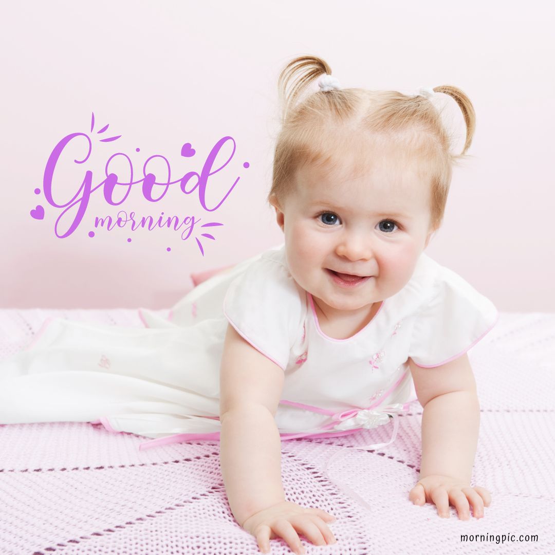 A cheerful baby girl with light hair in pigtails, wearing a white dress, is crawling on a pink knitted blanket. The text "Good morning" in purple, decorative font is displayed to the left. The baby smiles at the camera against a pale pink background—perfect for Good Morning Baby Images.