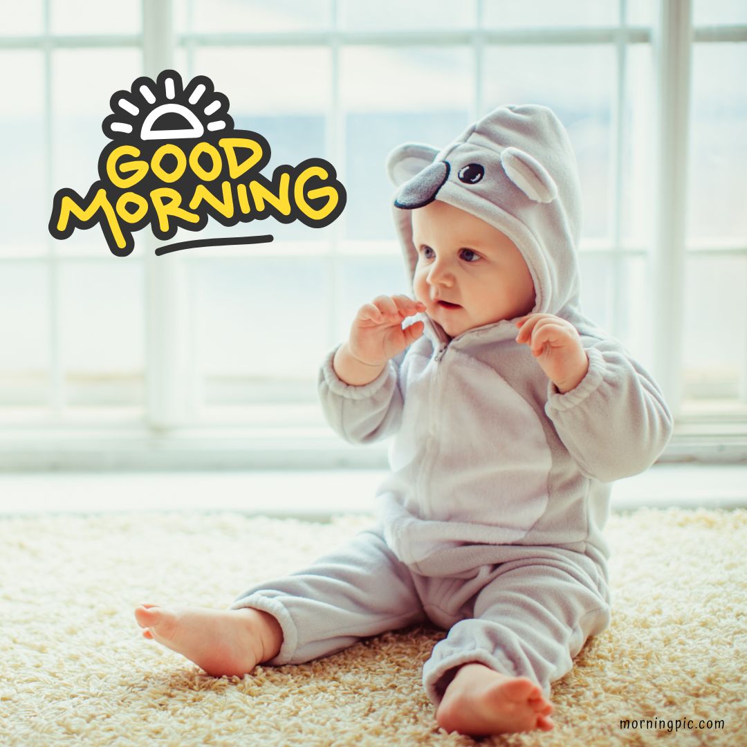 A baby dressed in a cozy gray bear-themed onesie sits on a light-colored carpet in front of a window. The image features the text "Good Morning" with a sun illustration