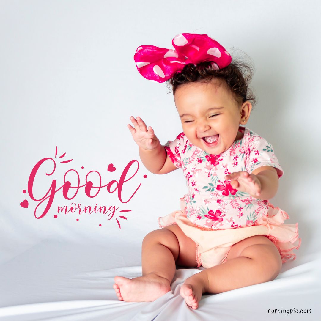 A smiling baby girl wearing a floral shirt and peach-colored shorts sits with hands raised against a white background. She has a large pink polka-dot bow on her head. The words "Good morning" are written in pink cursive font next to her, making it a perfect Good Morning Baby Girl image.