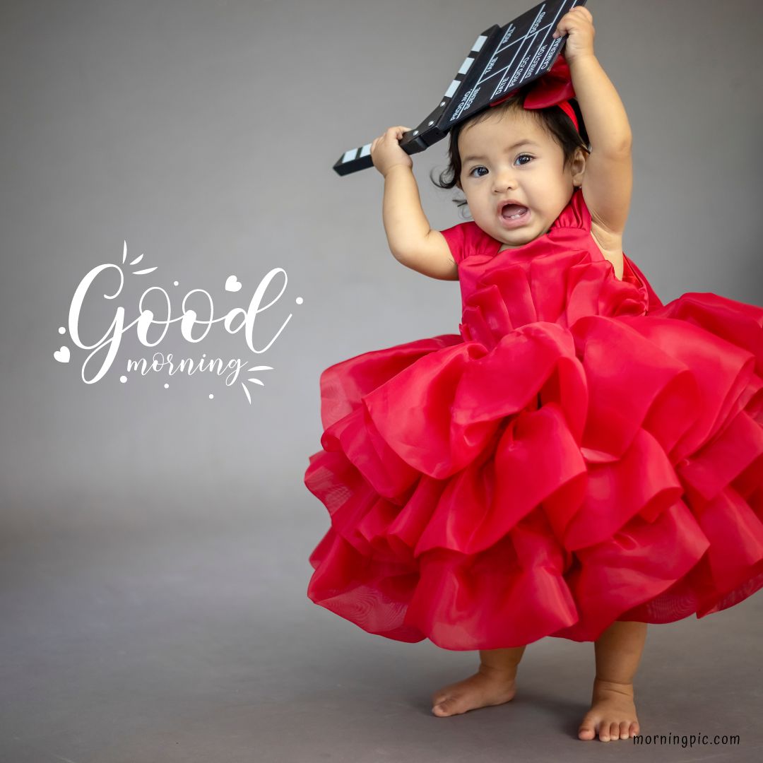 A smiling toddler wearing a frilly red dress holds a film clapperboard over her head. The words "Good morning" are written in cursive on the left side of the image. This adorable scene is perfect for Good Morning Baby Images with Girl Child Smiling. The background is plain gray.