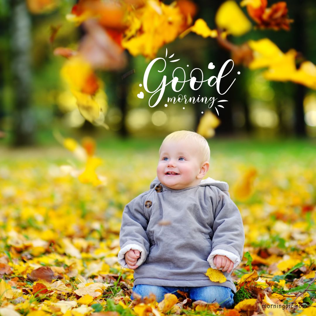 A happy baby wearing a gray coat sits on the ground surrounded by fallen autumn leaves. Bright yellow leaves are falling around them. The text "Good Morning" is written above the baby in a cheerful font. Perfect for those searching for Good Morning Baby Images HD, this scene captures pure joy and seasonal beauty.