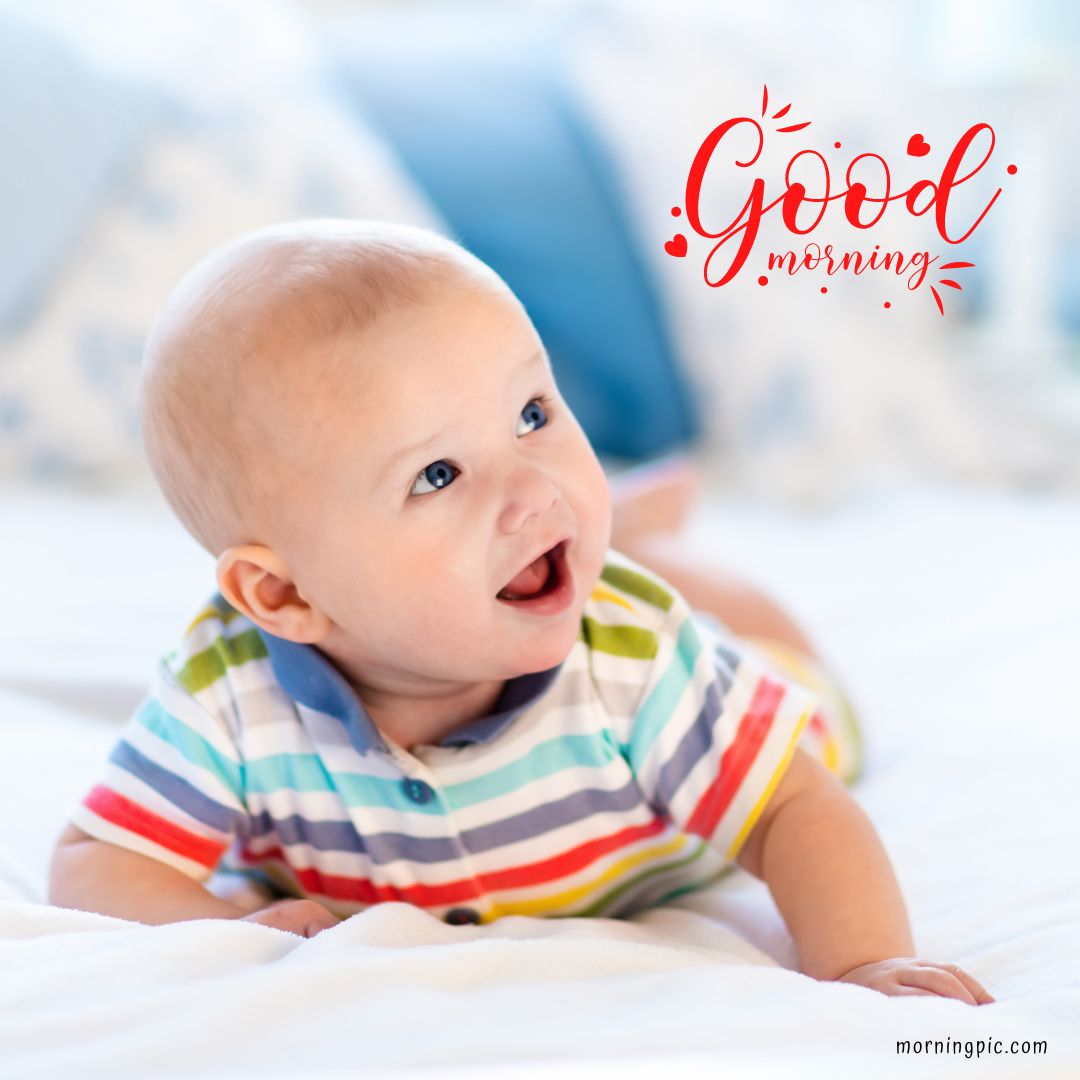 A smiling baby in a striped onesie lies on a white bed, looking up. The text "Good morning" is written in cheerful red letters on the top right corner of the image. This delightful scene exemplifies Good Morning Baby Images HD.