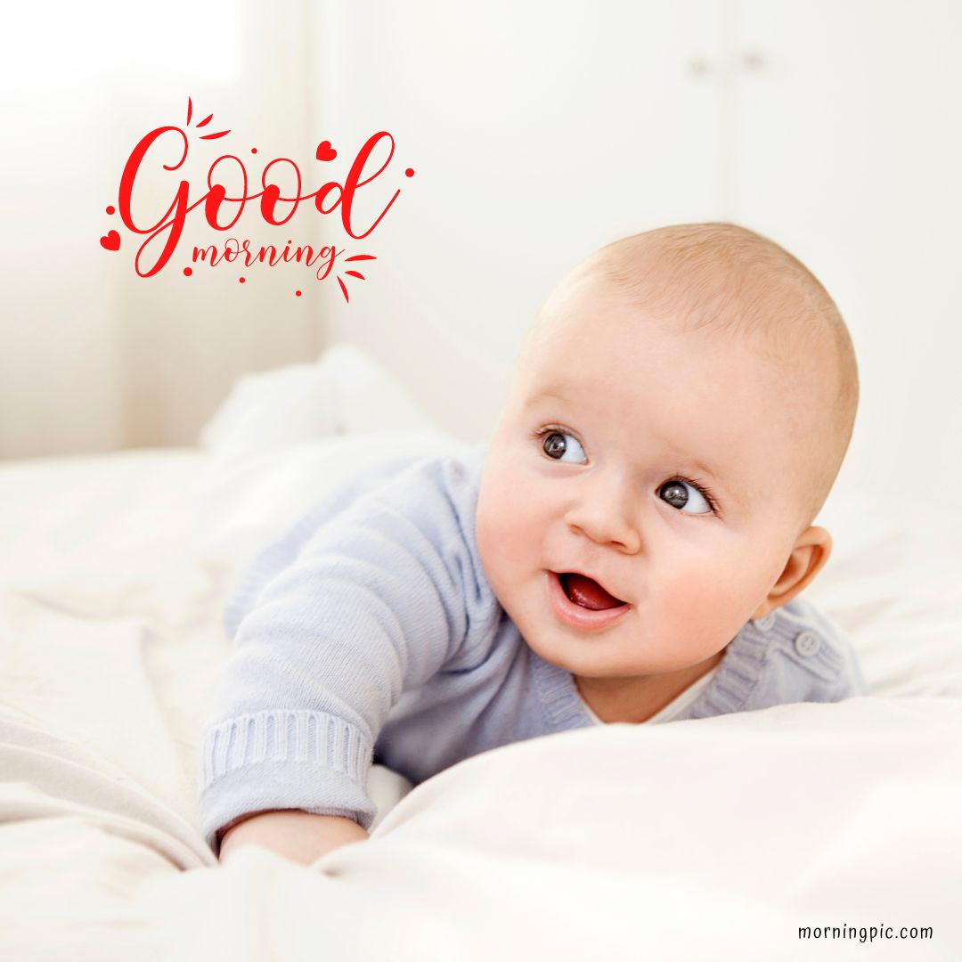 A smiling baby in a light blue sweater lies on their stomach on a white bed. The text "Good Morning" is written in red with small heart accents in the top left corner.