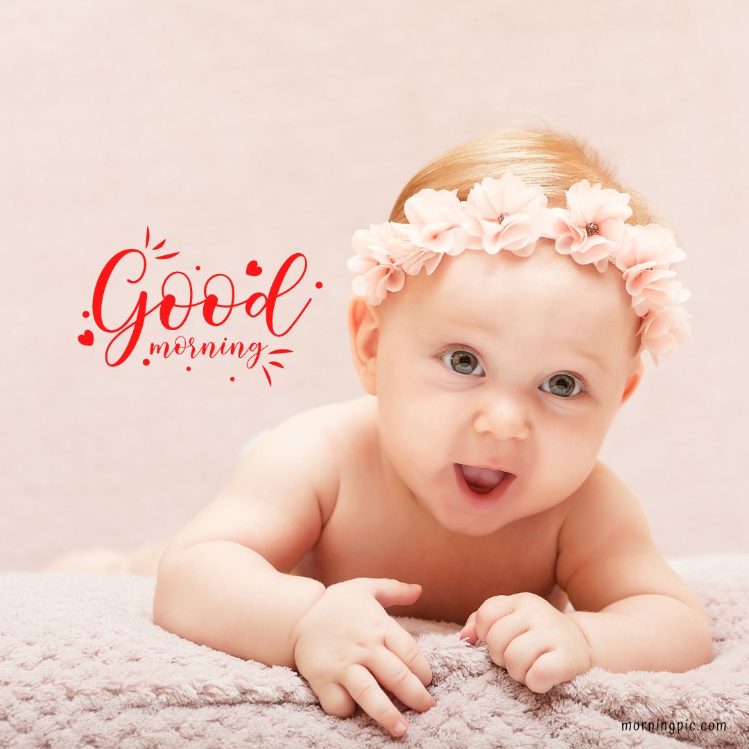 A baby girl lies on her stomach on a soft blanket, wearing a flower headband. She appears to be smiling or making a playful expression. The image includes the text "Good morning" in red letters with hearts and decorative lines around it, perfect for Good Morning Baby Girl images.