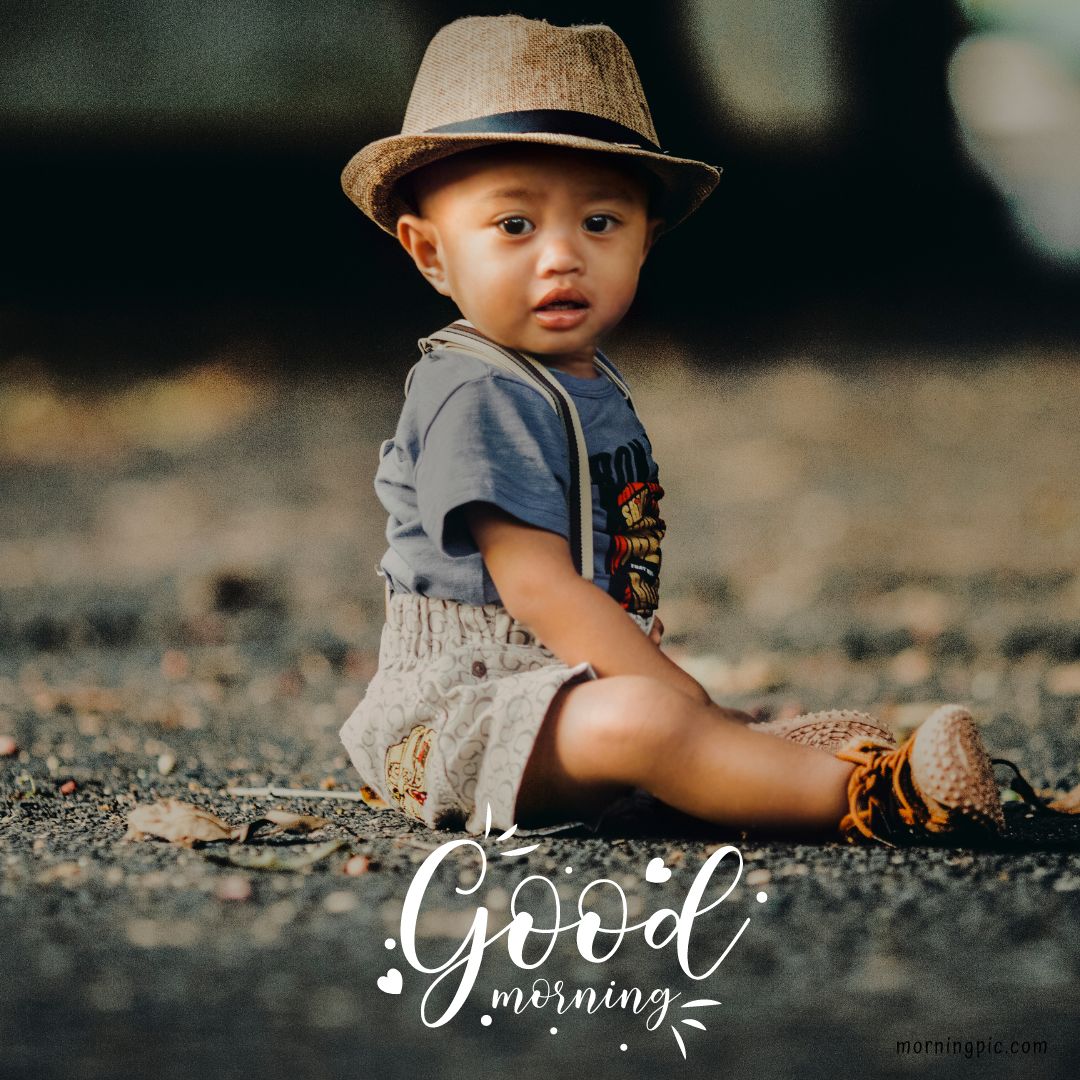 A young child wearing a beige hat sits on the ground, smiling in a blue shirt, patterned shorts, suspenders, and brown shoes. The background is blurred. The words "Good morning" are written in white with decorative elements near the bottom of the image. Perfect for Good Morning Baby Images HD.