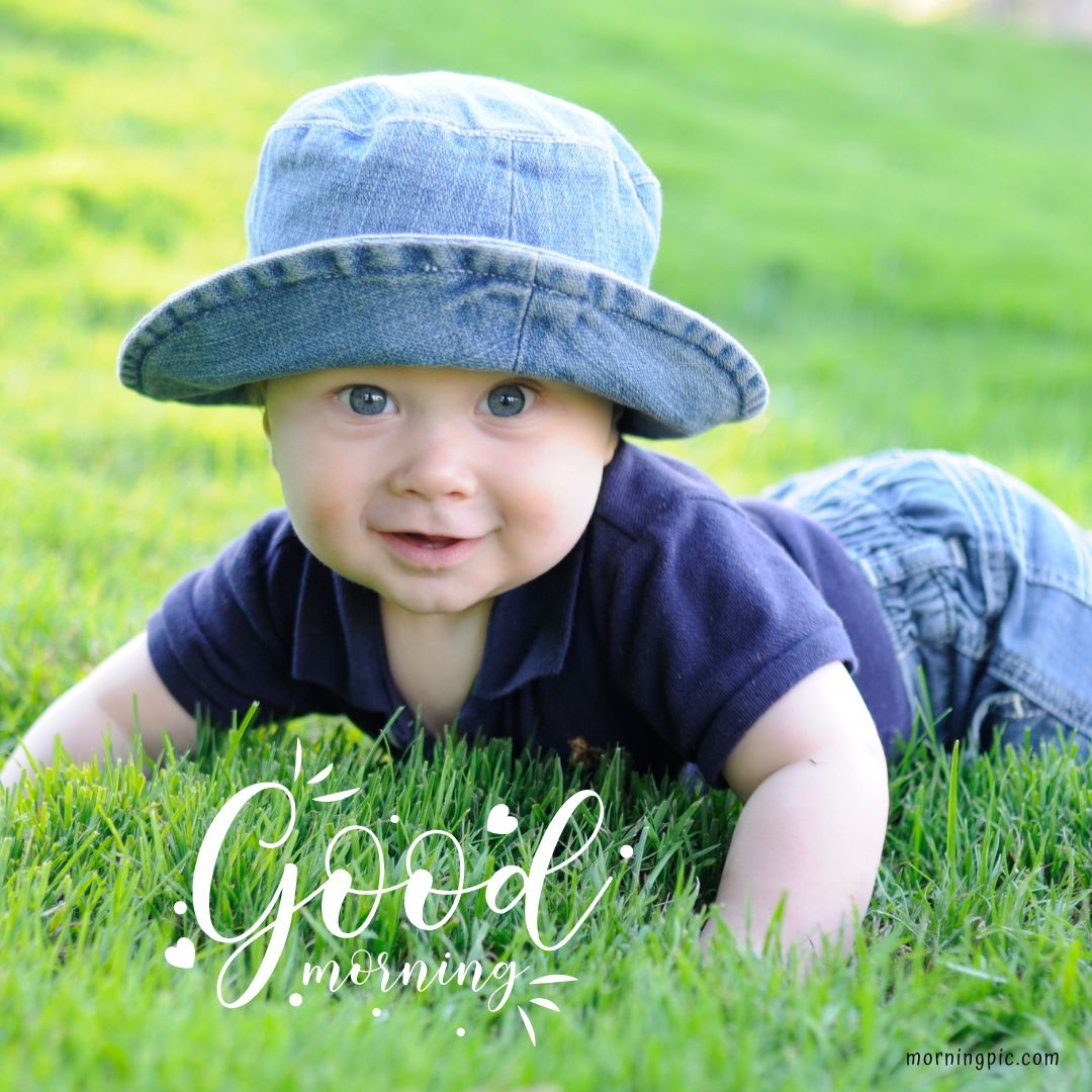 A baby boy wearing a blue hat and a navy blue shirt is crawling on green grass, smiling. The text "Good morning" is written in stylized white script at the bottom of the image. The background is a sunny, green outdoor area. Good morning images with baby boys capture pure joy!