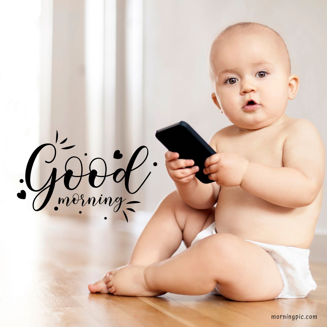 A baby girl wearing a diaper sits on a wooden floor holding a smartphone. She looks towards the camera with a neutral expression. The words "Good Morning" are written in stylish black cursive on the left side of the image.