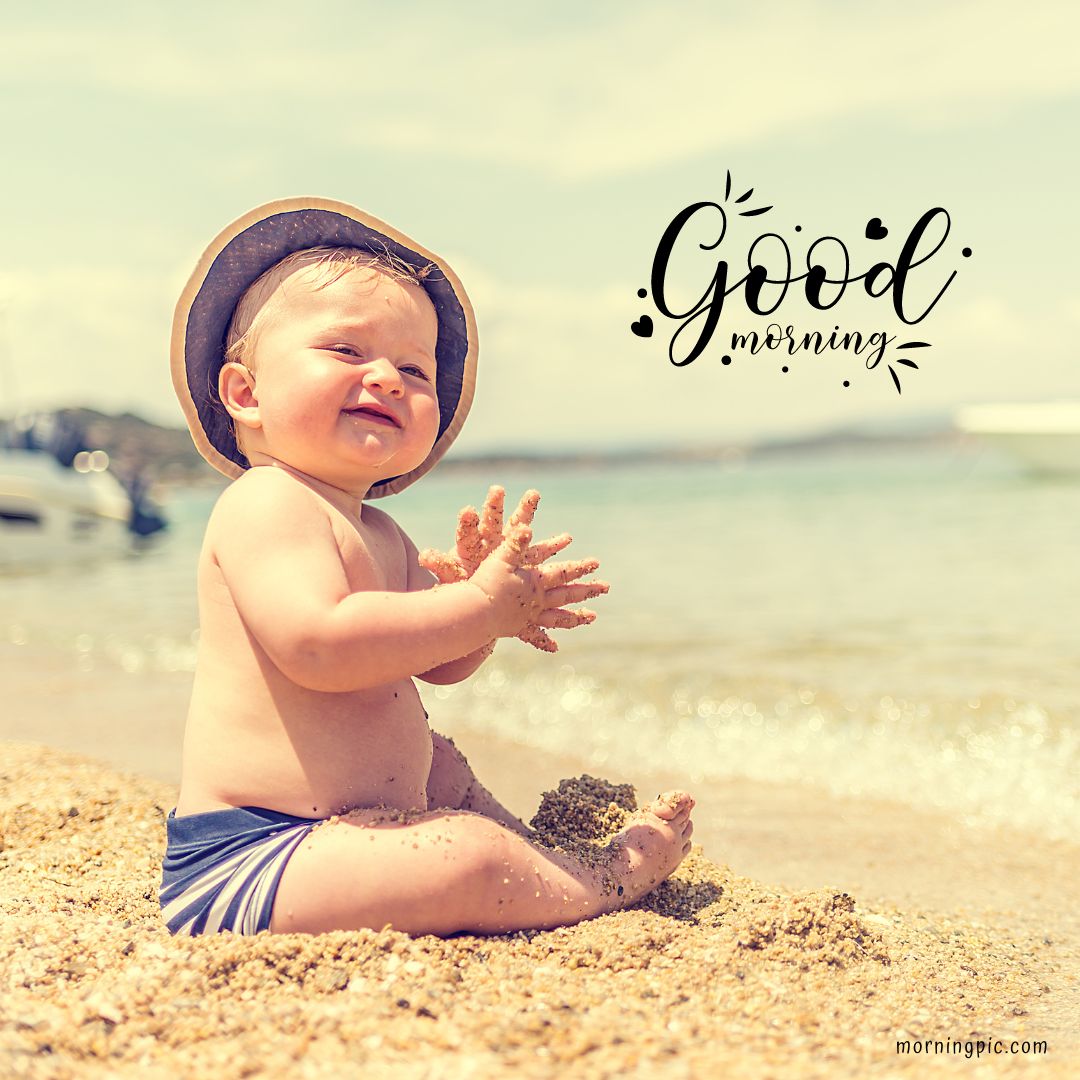 A smiling baby girl wearing a sun hat sits in the sand at a beach, clapping sandy hands with the sea and a boat in the background. The text "Good Morning Baby Girl" is written in decorative script in the sky above.