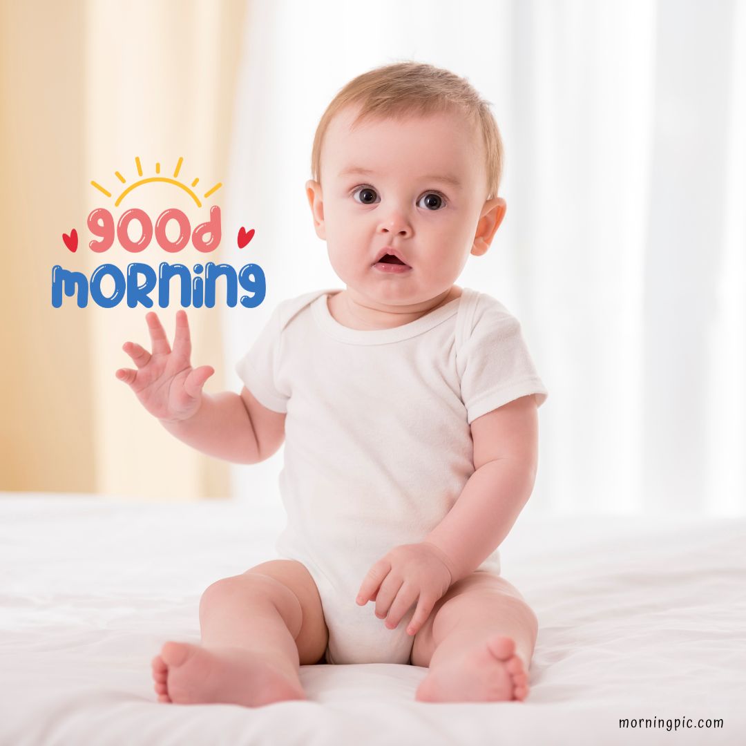 A baby with short light brown hair sits on a bed wearing a white onesie. The baby has a surprised expression and is waving one hand. The words "Good Morning" with a sun and hearts are written in colorful text next to the baby's head, perfect for good morning baby images.