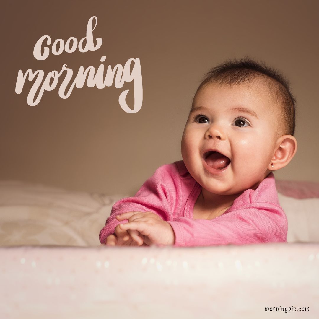 A baby wearing a pink outfit smiles joyfully while looking slightly to the side. The background is plain and light-colored. "Good morning" is written in cursive text in the top left corner, with "Good Morning Baby Images" 