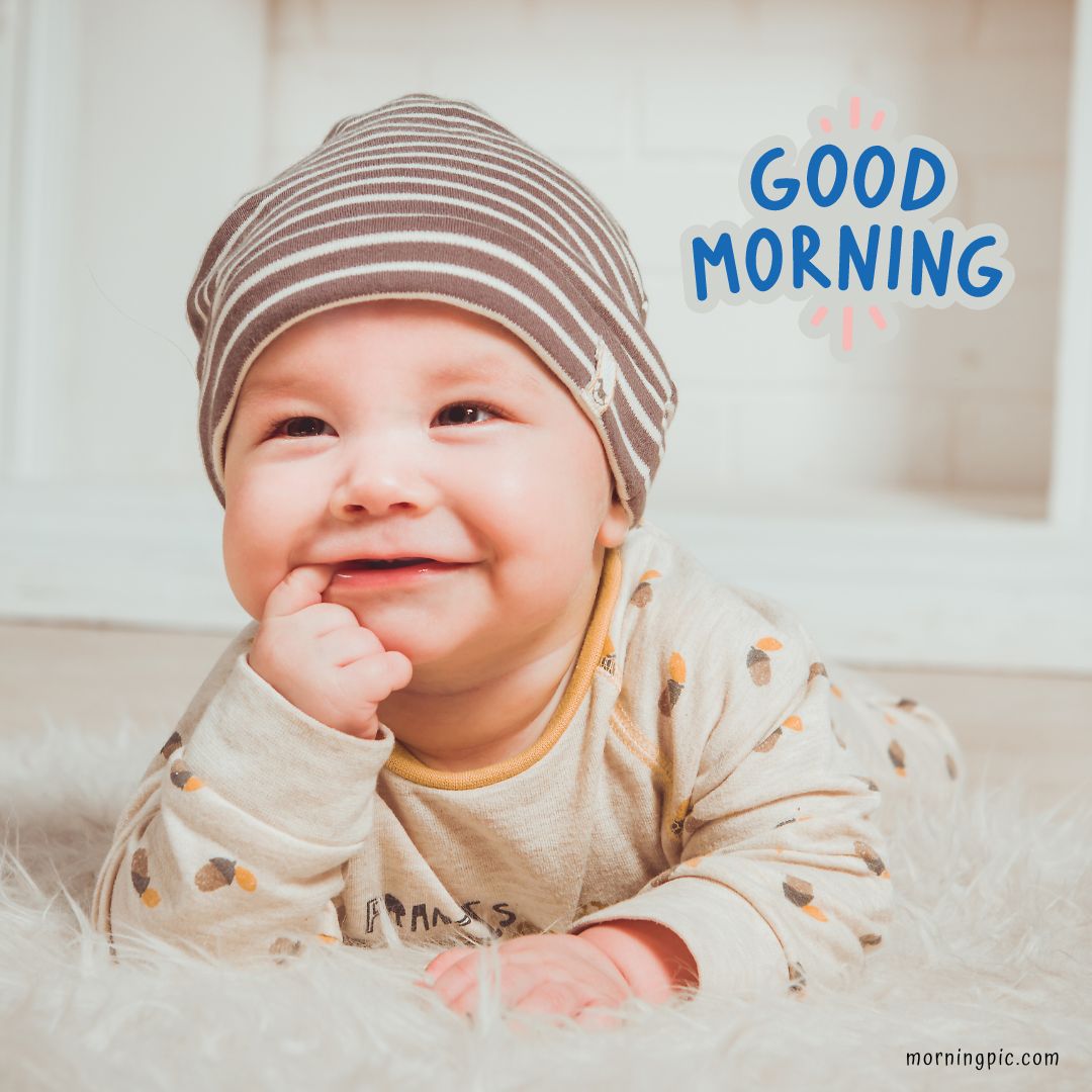 A smiling baby wearing a striped hat and patterned onesie lies on a soft, fluffy rug. The baby has their hand near their mouth and looks cheerful. The text "Good Morning Baby Images" is written in blue with red accents in the upper right corner.