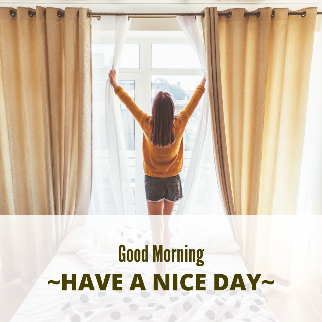 A person stands on a bed, stretching their arms towards a sunlit window with beige curtains. Wearing a yellow sweater and gray shorts, they are facing away from the camera. Text in the image reads: "Good Morning ~HAVE A NICE DAY~.