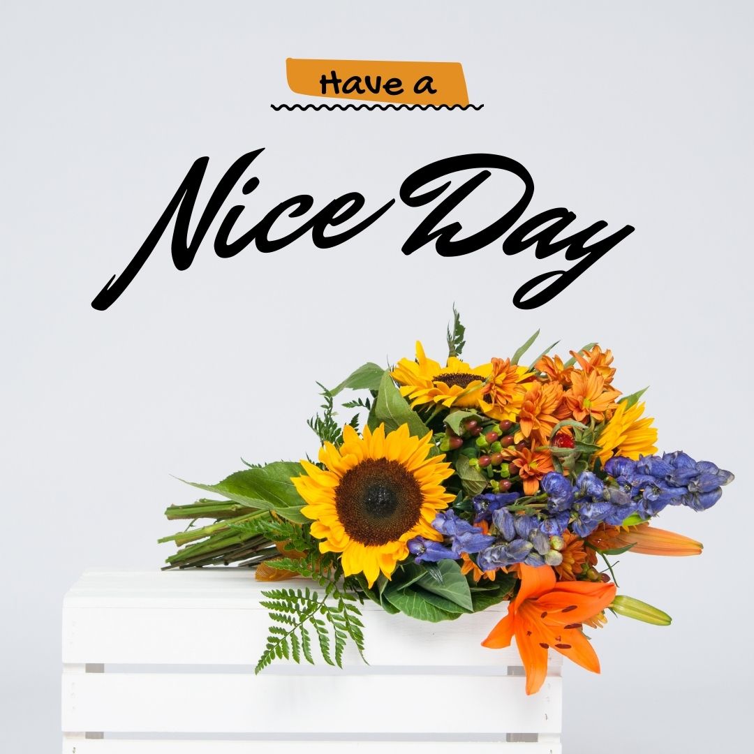 A colorful bouquet featuring sunflowers, orange lilies, and purple flowers sits atop a white wooden box. Above the bouquet, text reads "Good Morning Have a Nice Day" in black and orange font. The background is plain light gray.