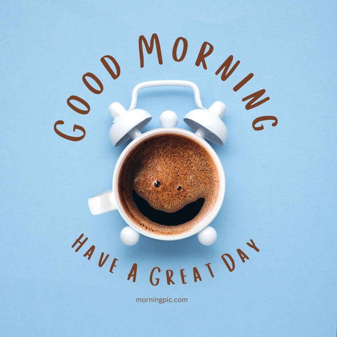 A cup of coffee with a smiley face made from foam on its surface, arranged to appear as an alarm clock with white bells and handle on a light blue background. Text curving around the cup reads, "Good Morning" at the top and "Have a Nice Day" at the bottom. It’s one of those images that instantly brighten your morning!