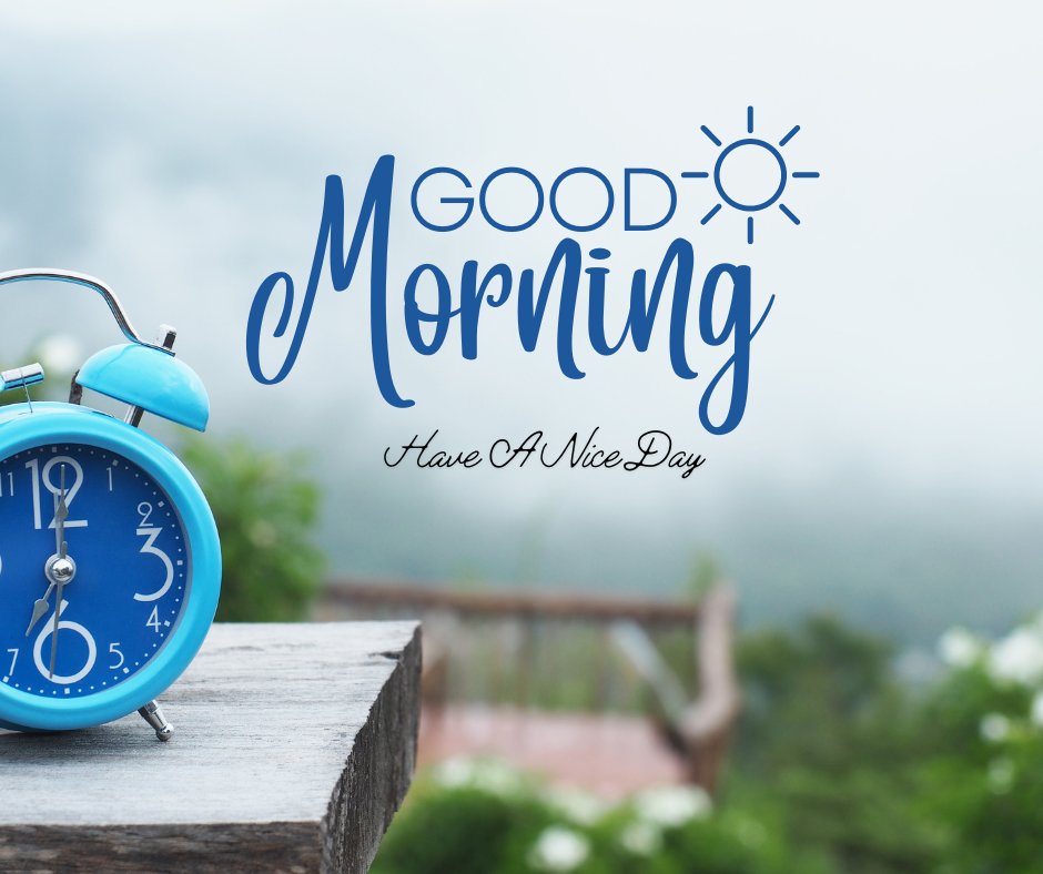 A blue alarm clock sits on a wooden table at the edge of a misty, green landscape. The words "GOOD Morning" are written in large, stylish blue letters with a small sun illustration. Below it, in elegant script, the phrase "Have A Nice Day" completes this picturesque scene reminiscent of good morning have a nice day images.