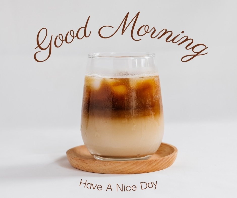 A glass of iced coffee with a layer of milk at the bottom, placed on a wooden coaster against a plain white background. The text "Good Morning" curves above the glass, and "Have A Nice Day" is written below, capturing the perfect morning vibe in vibrant images.