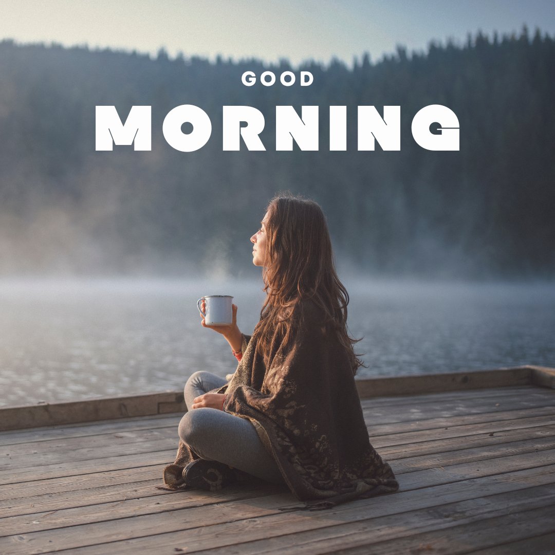 Good morning nature image of a young woman sitting cross-legged on a wooden dock, enjoying a misty lake view at sunrise, warmly wrapped in a blanket while holding a steaming mug.