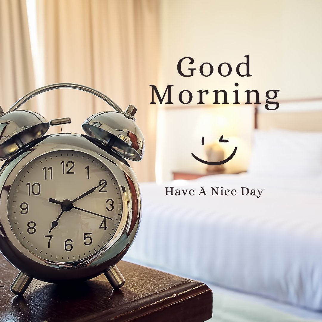 A silver alarm clock displays 7:00 and sits on a wooden nightstand. Behind it is a neatly made bed with white linens and a lit lamp. The image greets you with the words "Good Morning" and "Have A Nice Day," accompanied by a smiling face.